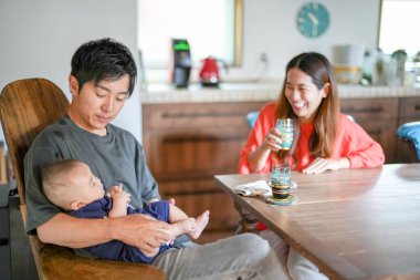 A Japanese father in his 30s holds a three-month-old baby while sitting at a Mediterranean-style dining table, sharing coffee and conversation with the mother in her 20s. Summer. Japan.