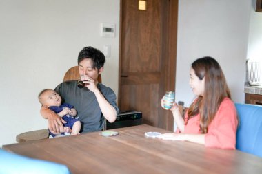 A Japanese father in his 30s holds a three-month-old baby while sitting at a Mediterranean-style dining table, sharing coffee and conversation with the mother in her 20s. Summer. Japan.