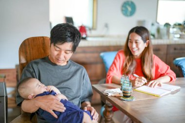A man is holding a baby in his arms while sitting at a table with a woman and a clock in the background. The woman is smiling and the man is looking at the baby