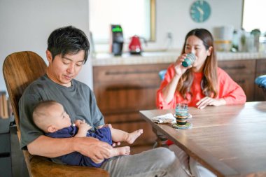 A Japanese father in his 30s holds a three-month-old baby while sitting at a Mediterranean-style dining table, sharing coffee and conversation with the mother in her 20s. Summer. Japan.