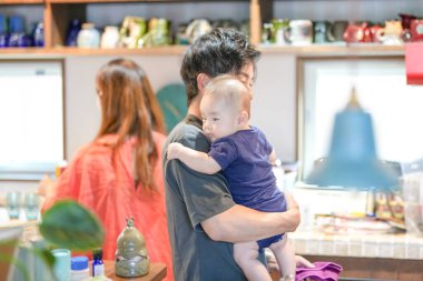 In a Mediterranean-style dining kitchen, a Japanese father in his 30s holds a three-month-old baby while wiping the table and talking to the mother in her 20s wearing orange clothes. Summer. Japan.