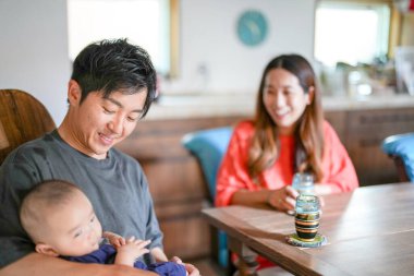 A Japanese father in his 30s holds a three-month-old baby while sitting at a Mediterranean-style dining table, sharing coffee and conversation with the mother in her 20s. Summer. Japan.