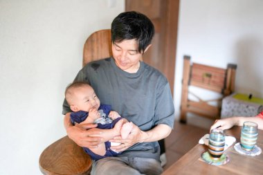 A Japanese father in his 30s holds a three-month-old baby while sitting at a Mediterranean-style dining table, sharing coffee and conversation with the mother in her 20s. Summer. Japan.
