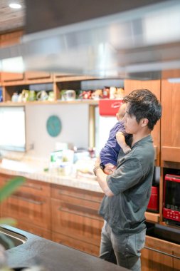 In a Mediterranean-style dining kitchen, a Japanese father in his 30s holds a three-month-old baby while wiping the table and talking to the mother in her 20s wearing orange clothes. Summer. Japan.