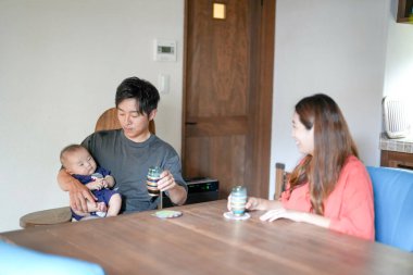 A Japanese father in his 30s holds a three-month-old baby while sitting at a Mediterranean-style dining table, sharing coffee and conversation with the mother in her 20s. Summer. Japan.