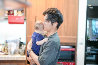 In a Mediterranean-style dining kitchen, a Japanese father in his 30s stands holding a three-month-old baby. Summer. Japan.