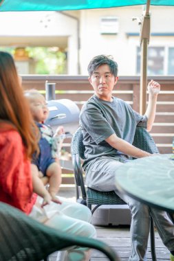 A man sits at a table with two other people, one of whom is holding a baby. The man is wearing a gray shirt and he is looking at something. The scene is casual and relaxed