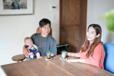 A Japanese father in his 30s holds a three-month-old baby while sitting at a Mediterranean-style dining table, sharing coffee and conversation with the mother in her 20s. Summer. Japan.