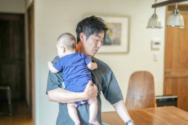In a Mediterranean-style living room, a Japanese man in his 30s wipes the table with a cloth while holding a 3-month-old baby. Summer. Japan.
