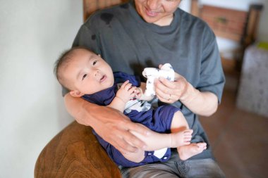 A Japanese father in his 30s holds a three-month-old baby while sitting at a Mediterranean-style dining table, sharing coffee and conversation with the mother in her 20s. Summer. Japan.