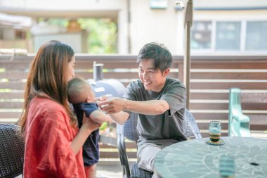 A man is holding a baby while a woman sits behind him. The baby is smiling and the man is smiling as well. The scene is happy and warm, with the family enjoying each other's company