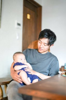A man is holding a baby in his arms. The baby is smiling and the man is looking at the camera. The scene is warm and loving, with the man and baby sharing a special moment together