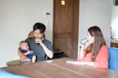 A Japanese father in his 30s holds a three-month-old baby while sitting at a Mediterranean-style dining table, sharing coffee and conversation with the mother in her 20s. Summer. Japan.
