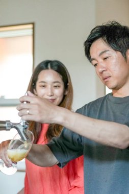 A man and a woman are standing next to a tap, with the man pouring a drink into a glass. The woman is watching him, and the atmosphere seems to be casual and relaxed