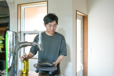 A man is standing in a kitchen, holding a bottle of olive oil and a plate. He is preparing to cook something