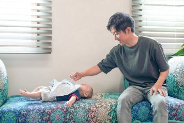 A man is holding a baby on a couch. The baby is wearing a blue shirt and a blanket