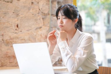 A woman is sitting at a table with a laptop in front of her. She is wearing a white shirt and has her hair in a ponytail. She is focused on her work, possibly typing or browsing the internet