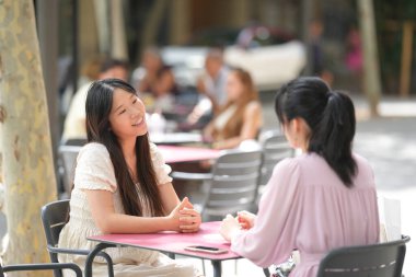 Two women sitting at a table in a park, one of them smiling. The other woman is looking at her phone. There are other people in the background, some of them sitting at tables and others standing
