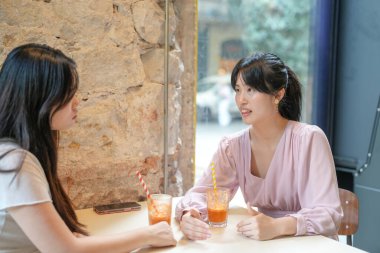 Two women are sitting at a table in a cafe, talking and drinking juice. One of the women is holding a cell phone
