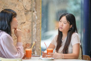 Two women are sitting at a table with orange juice and straws. One of them is looking at the other