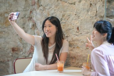 Two women sitting at a table, one taking a picture of the other. The woman taking the picture is smiling and the other woman is also smiling
