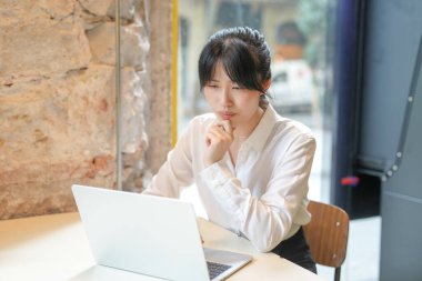 A woman is sitting at a table with a laptop in front of her. She is looking at the screen and she is deep in thought