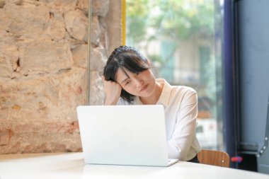A woman is sitting at a table with a laptop in front of her. She is tired and is leaning her head on the laptop
