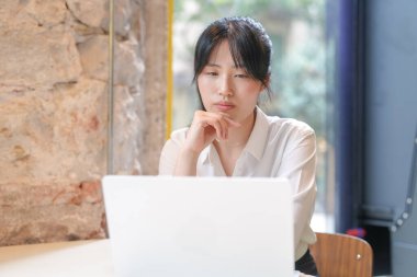 A woman is sitting at a table with a laptop open in front of her. She is looking at the screen with a thoughtful expression on her face