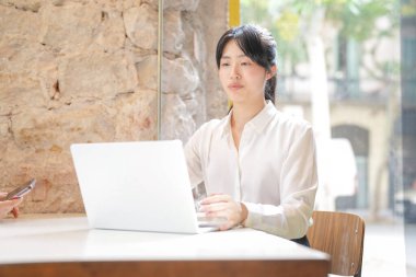 A woman is sitting at a table with a laptop in front of her. She is wearing a white shirt and she is focused on her work