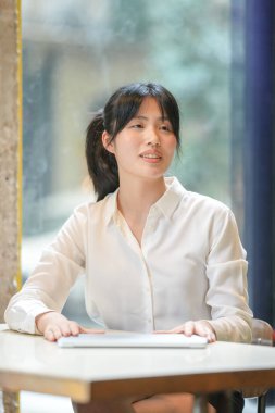 A woman is sitting at a table with a laptop and a book. She is wearing a white shirt and has her hair in a ponytail. The scene suggests that she is working or studying, possibly for a class or a job