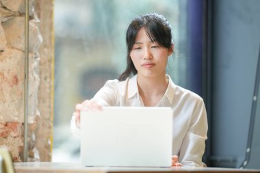A woman is sitting at a table with a laptop in front of her. She is looking at the screen with a frown on her face
