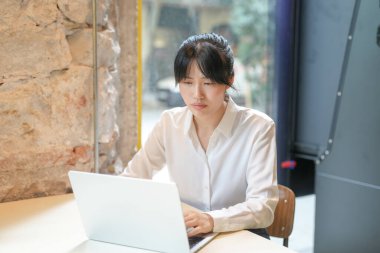A woman is sitting at a table with a laptop in front of her. She is wearing a white shirt and has her hair in a bun. She is focused on her work, possibly typing or browsing the internet