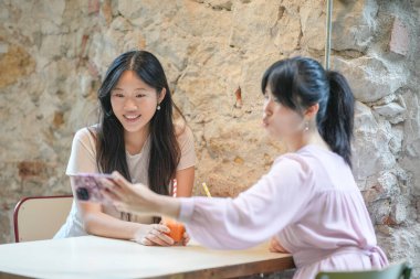 Two women sitting at a table, one of them taking a picture of the other with a cell phone