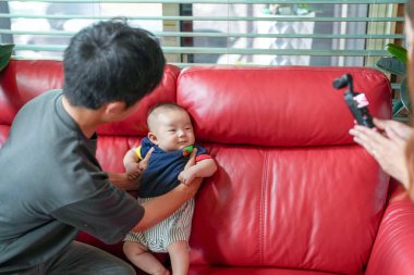 A man is holding a baby on a red couch. The baby is wearing a blue shirt and a striped shirt