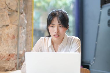 A woman is sitting at a table with a laptop in front of her. She is wearing a white shirt and has her hair in a ponytail. Concept of focus and concentration as the woman works on her laptop