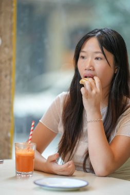 A woman is eating a sandwich while drinking a glass of orange juice. She is looking at the camera