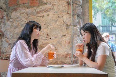 Two women are sitting at a table, drinking juice and talking. Scene is friendly and relaxed