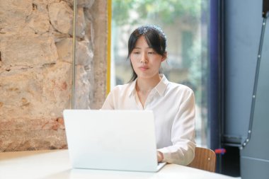 A woman is sitting at a table with a laptop in front of her. She is wearing a white shirt and has her hair in a ponytail. Concept of focus and concentration as the woman works on her laptop