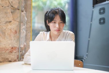 A woman is sitting at a table with a laptop in front of her. She is looking at the screen with a serious expression on her face