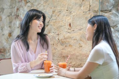 Two women are sitting at a table, drinking orange juice and talking. Scene is friendly and relaxed