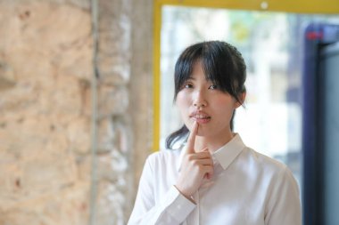 A woman with a white shirt and black hair is looking at the camera. She is holding her hand to her mouth and she is thinking