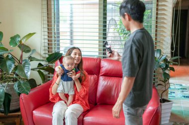 A woman is holding a baby on a red couch while a man takes a picture of them. The scene is warm and friendly, with the man capturing a special moment between the mother and child
