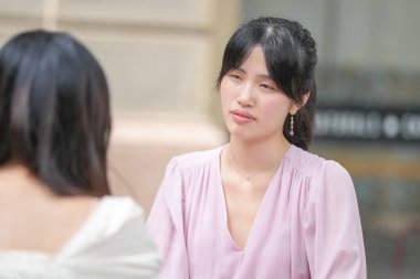 Two women are sitting at a table, one of them is wearing a pink shirt. The woman in the pink shirt is looking at the other woman, who is wearing a white shirt