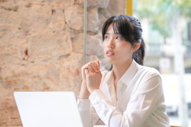 A woman is sitting at a table with a laptop in front of her. She is looking at the laptop with a serious expression on her face