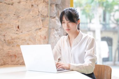 A woman is sitting at a table with a laptop in front of her. She is wearing a white shirt and has her hair in a ponytail. Concept of focus and concentration as the woman works on her laptop