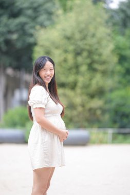 A woman is standing in a park, wearing a white dress and smiling. She is pregnant and she is enjoying her time outdoors