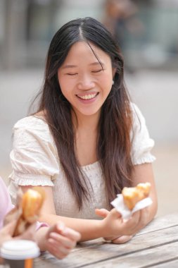 A woman is sitting at a table with a pastry in her hand. She is smiling and she is enjoying her food