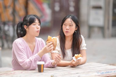 Two women are sitting at a table eating food. One of them is eating a hot dog. The other woman is eating a sandwich. They are both looking at something in the distance