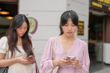 Two women are walking down the street, one of them is looking at her phone. The other woman is looking at her phone as well