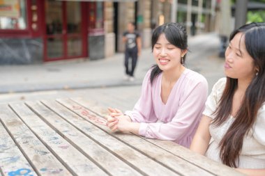 Two women sitting at a wooden table, one of them smiling. The other woman is looking at her with a serious expression. Scene is one of friendship and companionship